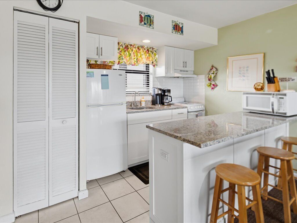 A kitchen with white cabinets, a central island with stools, a microwave, and a clock on the wall.
