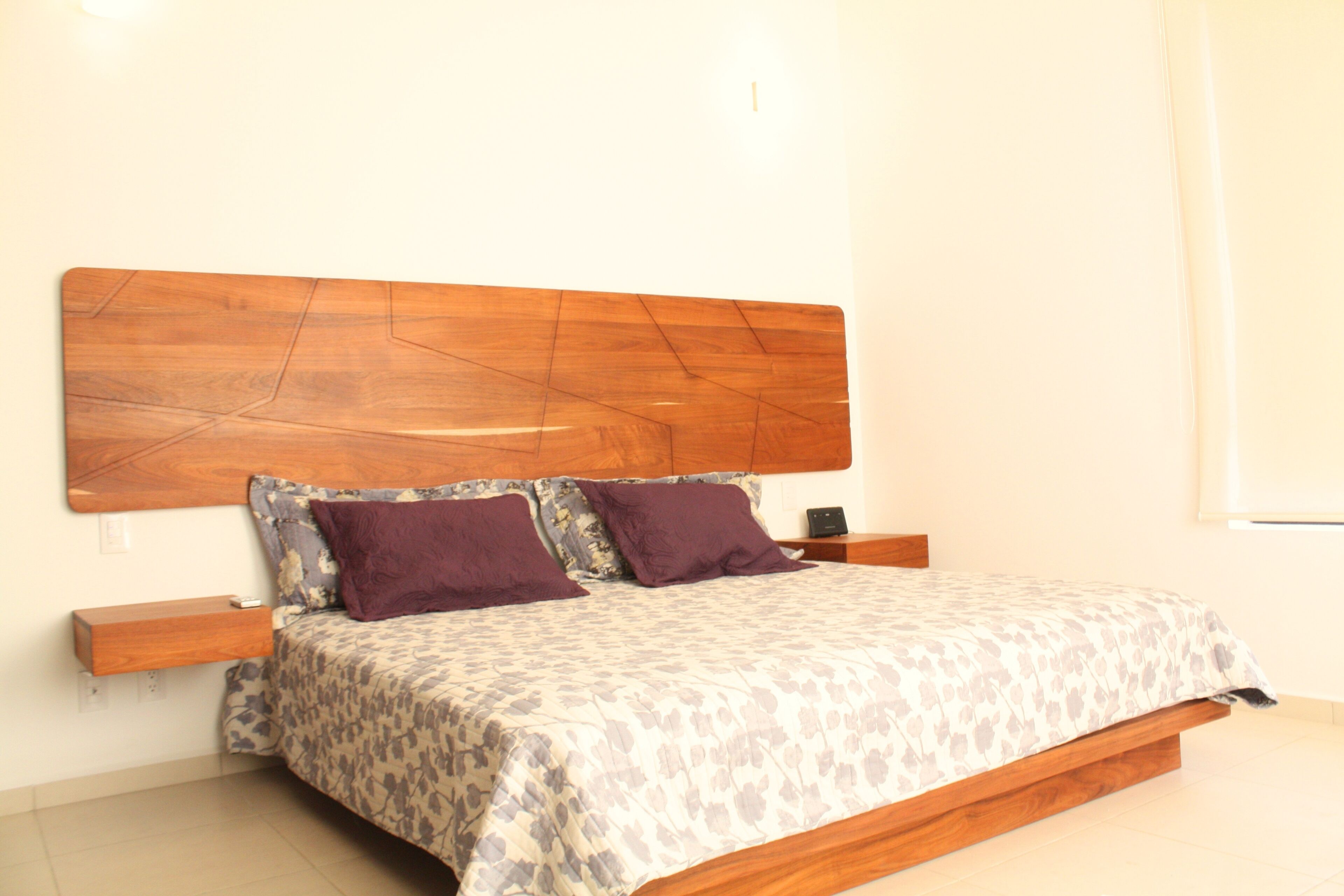 Un dormitorio con cabecera de madera, una cama con ropa de cama estampada, dos mesitas de noche y una pequeña estantería de madera.