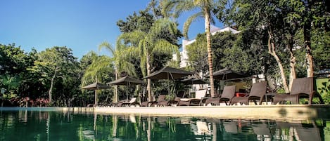 A poolside area with lounge chairs and umbrellas, surrounded by palm trees and a clear sky.