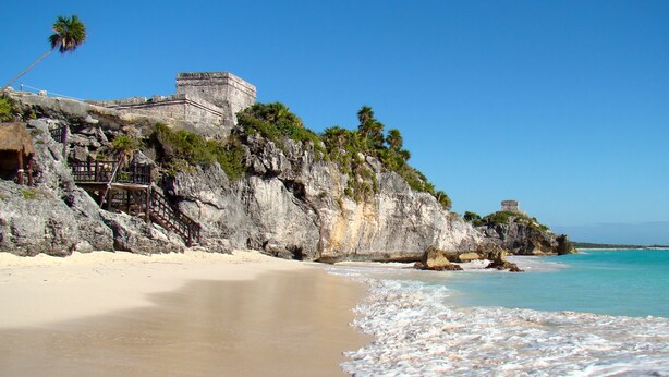 A beach with clear turquoise waters, a sandy shore, and a stone structure on a cliff.