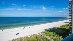 A beach view with a white sandy shore, clear blue water, and a building with balconies. - Beachfront 1BR Apollo condo with heated pool, WiFi, and Gulf views. (Marco Island)