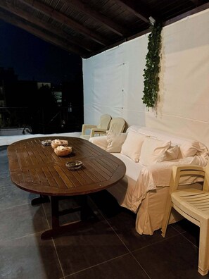An outdoor patio with a round wooden table, white cushioned seating, and a white wall with a hanging plant.