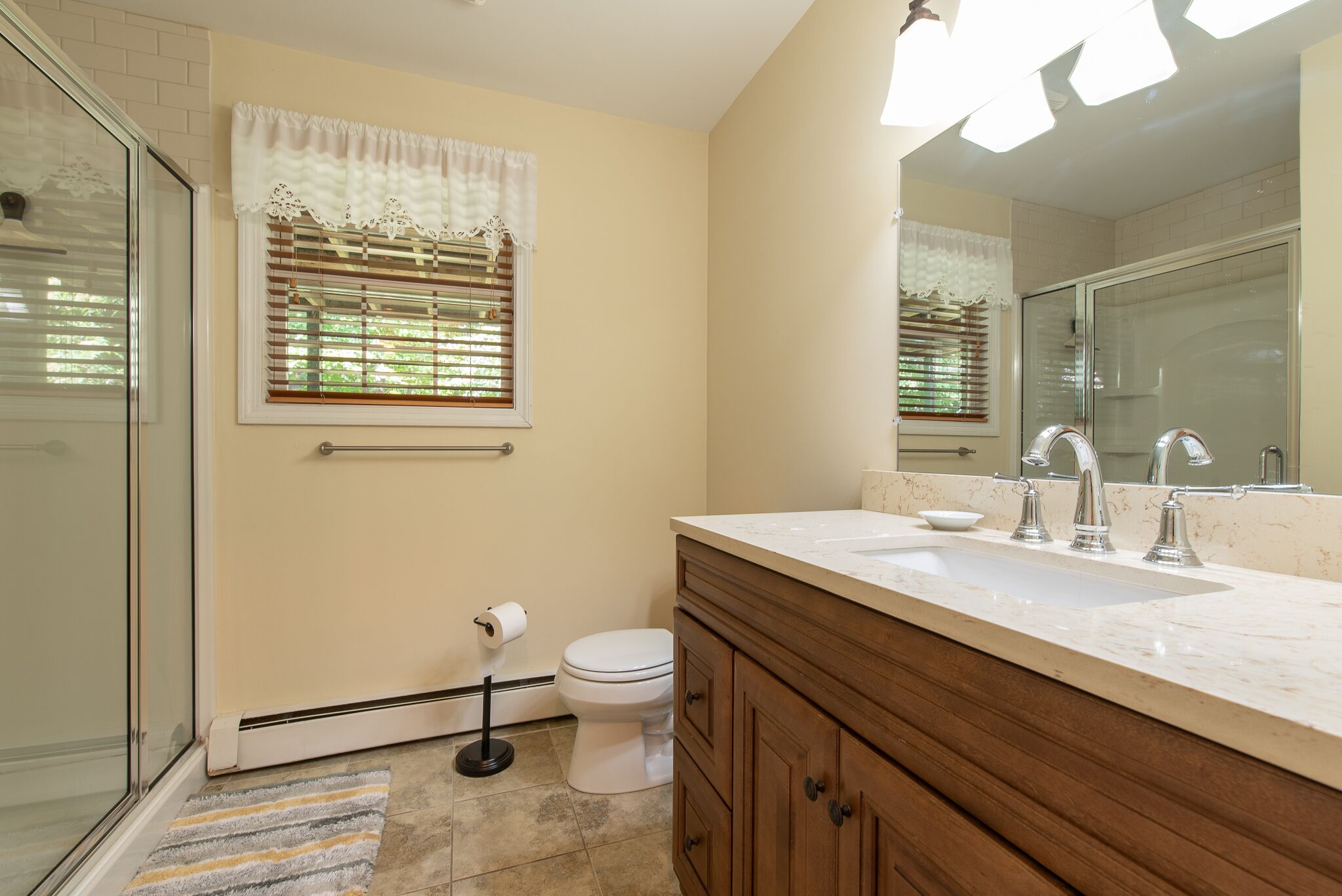 Une salle de bain avec une douche à parois de verre, des toilettes, un lavabo avec un meuble-vasque et une fenêtre avec des stores.