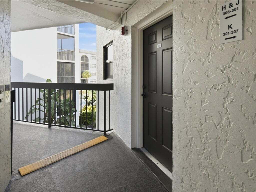 Entrada de un edificio con una reja metálica negra, una puerta oscura y una pared blanca con textura que muestra los números de apartamentos H & J 306-301 y K 301-307.