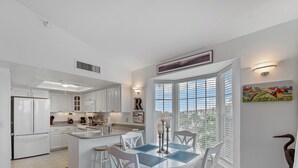 A modern kitchen with white cabinetry, a central island, and a dining area with a round table and chairs. - Enjoy a Beautiful Gulf Sunset from the Screened Porch on Resort Villa A3221B (Cape Haze)