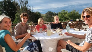Un groupe de personnes est réuni autour d’une table à l’extérieur, où elles prennent leur repas. La table est recouverte d’une nappe en dentelle blanche, et l’ensemble est entouré d’un paysage naturel.