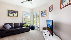 A living room with a dark brown leather sofa, a flat-screen TV on a white stand, and a large window with white blinds.