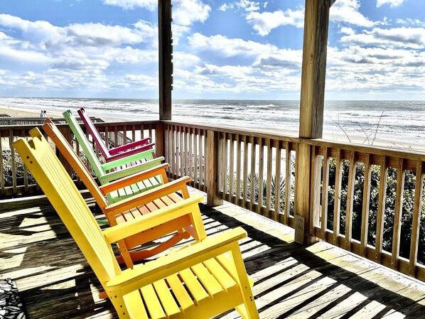 Um deck de madeira com cadeiras Adirondack coloridas com vista para uma praia.