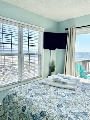 Quarto com cama, televisão, planta em vaso e vista para a praia.