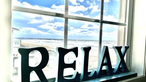 Ein Fenster, auf dem in großen Buchstaben das Wort 'RELAX' steht, mit Blick auf einen Strand, Wellen und einen klaren Himmel.