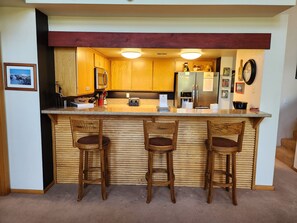 A kitchen with wooden cabinets, a stainless steel refrigerator, a built-in bar with three stools, and a clock on the wall. - Snowcreek Resort Vacation Rental 838 (Mammoth Lakes)