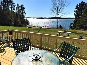 A deck with a glass table and chairs, overlooking a lake with chairs and a picnic table.