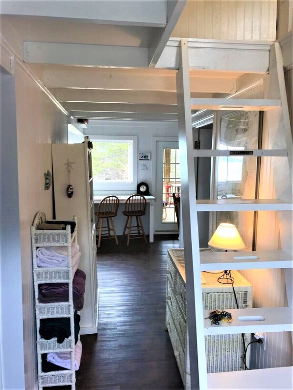 A narrow hallway with a staircase on the right, leading to a room with a lamp and a small table. A wicker basket rack is on the left, and a dining area with chairs and a table is visible in the background.