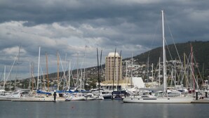 A marina with multiple sailboats docked, a large building in the background, and a cloudy sky.