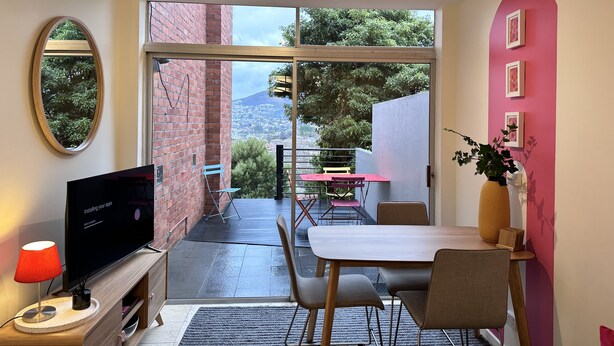 A modern room with a TV, a dining table, and chairs. There is a large window with a view of a cityscape and greenery.