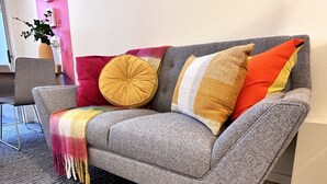 A grey sofa with a multicolored throw blanket, a round mustard-colored cushion, and two striped pillows in shades of white, yellow, and red.
