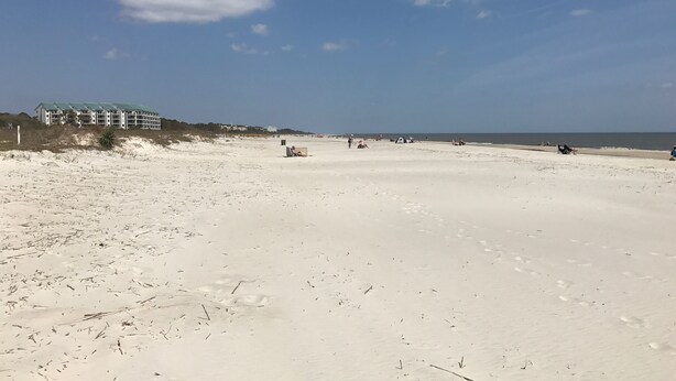 En bred, sandet strand med fodspor, et par mennesker og en bygning med grønt tag i baggrunden.