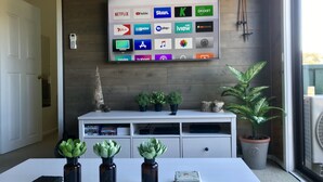 A modern living room with a flat-screen TV displaying various streaming services, a white entertainment unit with plants, and a large window with a view. - Yowie 3 - (Yowie Lodge) (Jindabyne)