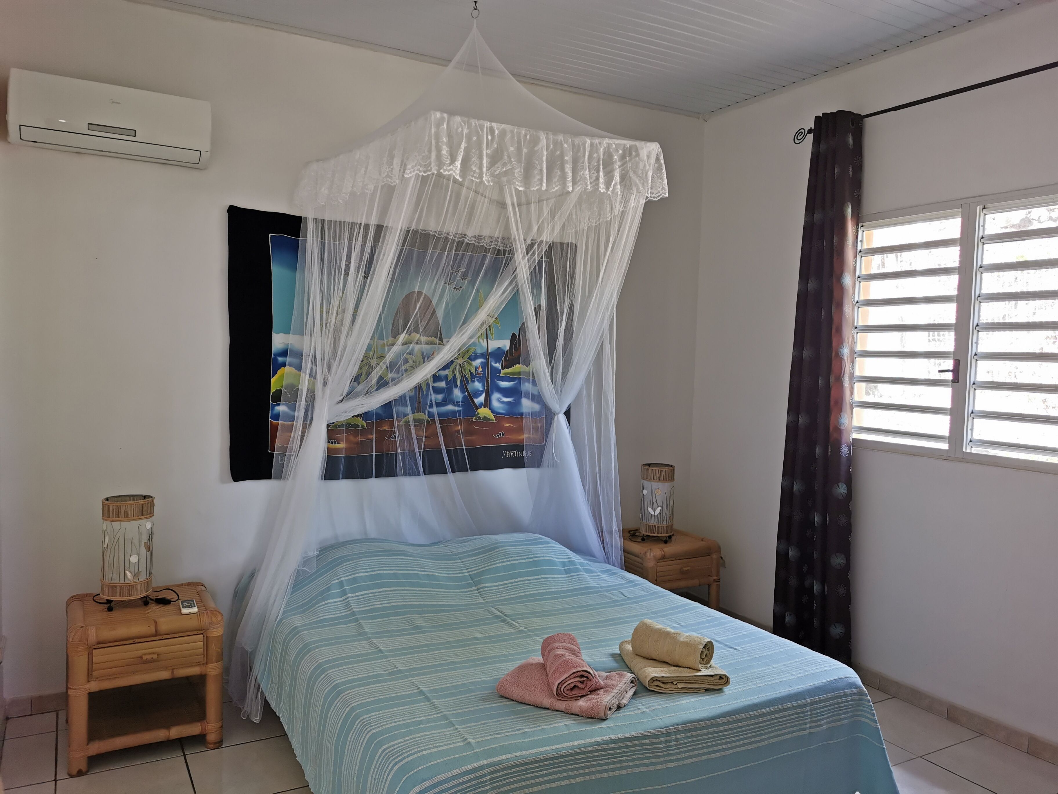 A bedroom with a bed, a mosquito net, a nightstand, a lamp, and a window with curtains.