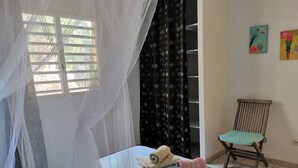 A room with a bed, a chair, a window with a curtain, and two wall-mounted paintings. - Hibiscus: calm and breathtaking views for this T3 overlooking the bay of Fort de France (Les Trois-Îlets)