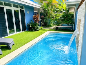 A swimming pool with a waterfall feature, surrounded by a well-maintained lawn and various tropical plants.