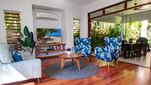 Una sala de estar moderna con un sofá blanco, una mesa de centro redonda de madera y dos sillones estampados azules. Se observa una estantería con libros y un cuadro con una escena de playa.