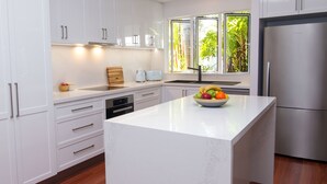 Una cocina moderna con muebles blancos, una isla grande, un refrigerador de acero inoxidable y una ventana con vista a follaje.