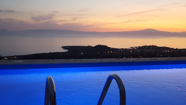 Una piscina con escaleras metálicas, vista a una costa distante y un atardecer de fondo.