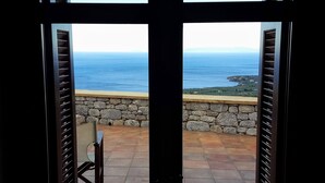Vista desde un balcón con paredes de piedra y una silla, con vistas a la costa, edificios y el mar.