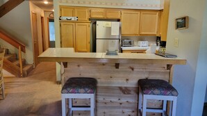 A kitchen with wooden cabinets, a refrigerator, a microwave, and a toaster. There are two bar stools with patterned cushions.