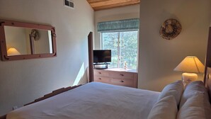 A bedroom with a large bed, a dresser, a television, and a window with a view of trees.