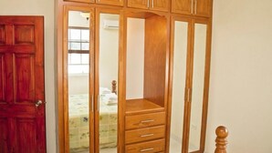 Une armoire en bois dotée de nombreux compartiments et de tiroirs, une porte en bois, et un lit avec une couvre-lit à motifs floraux en arrière-plan.