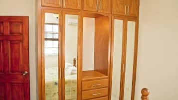 Un armario de madera con varios compartimentos y cajones, una puerta de madera, y una cama con colcha estampada floral al fondo.