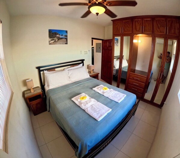 A hotel room with a bed, a nightstand, a bedside table, a ceiling fan, a wooden wardrobe, and a picture on the wall.