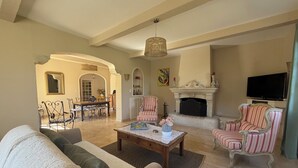 A spacious living room with a fireplace, striped armchairs, a coffee table, and a dining area with a chandelier.