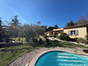 A pool with a stone border, a patio with chairs, and a building with a tiled roof.