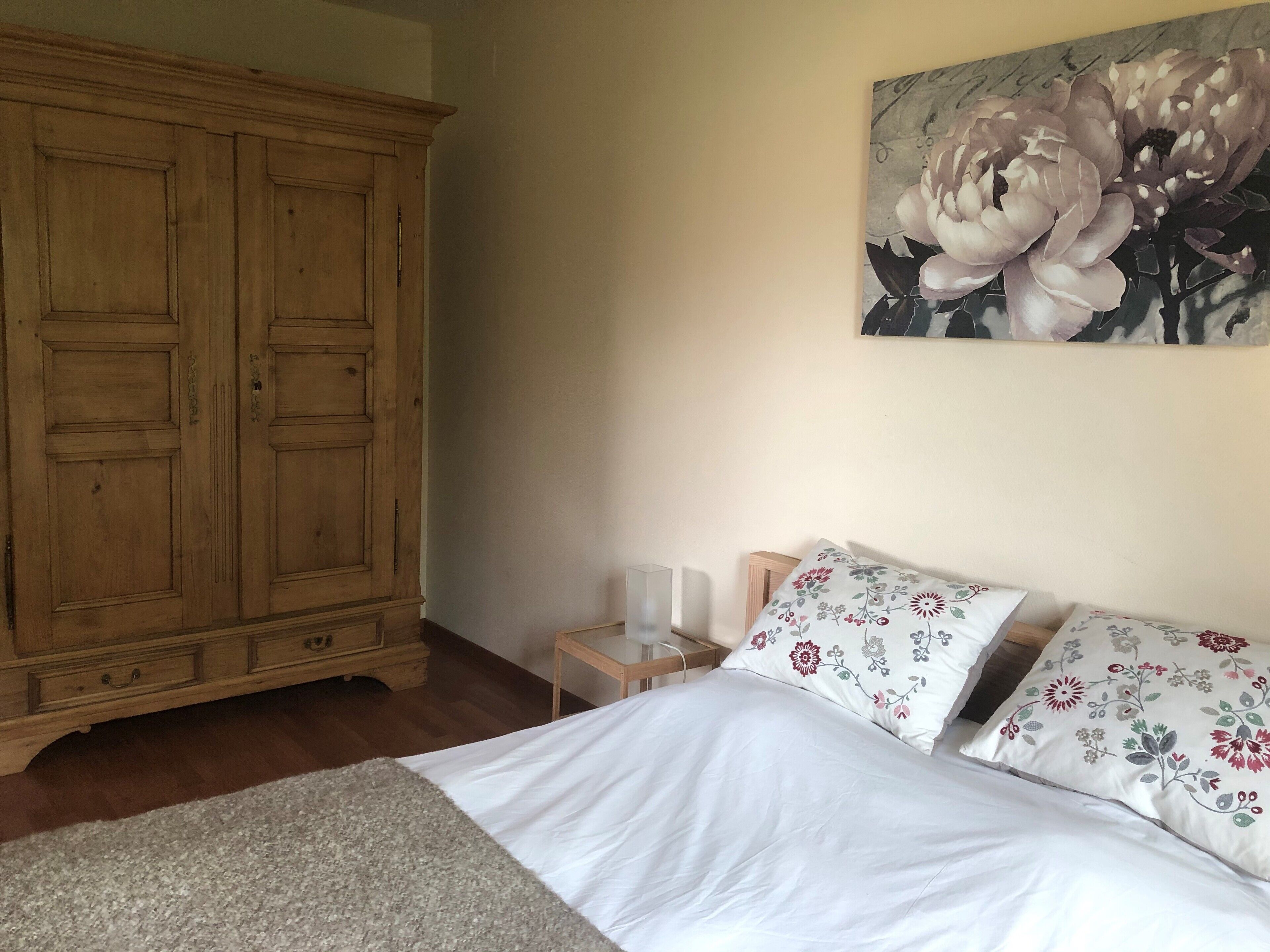 Une chambre avec une grande armoire en bois, un lit agrémenté d’oreillers à motifs floraux et un tableau représentant des fleurs accroché au mur.