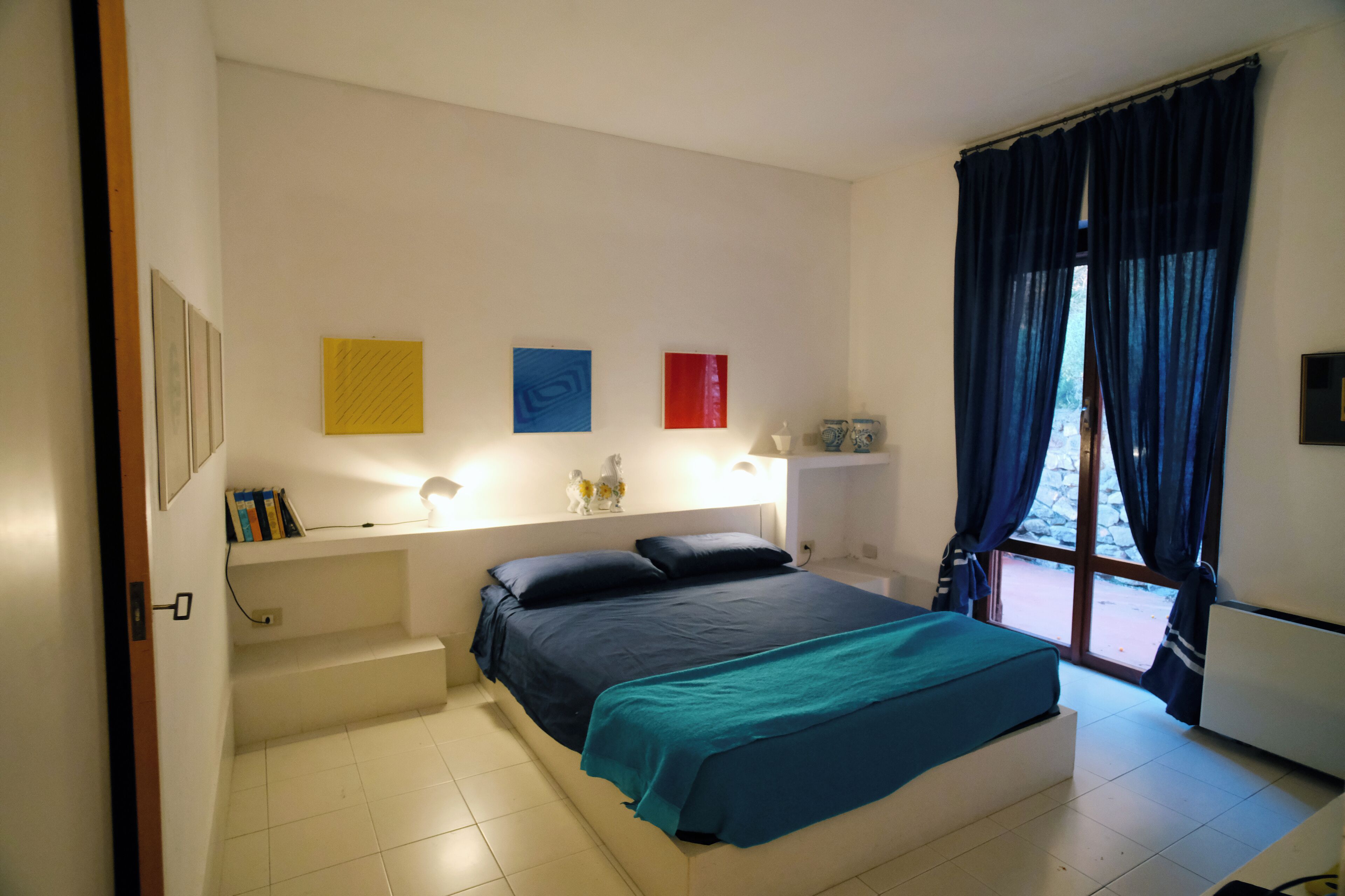 A bedroom with a bed, a nightstand, a window with curtains, and wall art.
