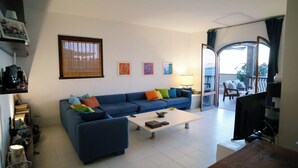 A living room with a blue sofa, a coffee table, and a TV. There is a balcony with chairs and a table. - Beautiful villa overlooking the sea in the Maremma National Park (Talamone)