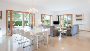 A spacious living area with a dining table, sofas, and a TV. Large windows offer views of greenery.