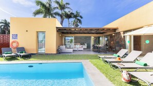 Un área de piscina con sillones reclinables, un patio cubierto y vistas a palmeras.