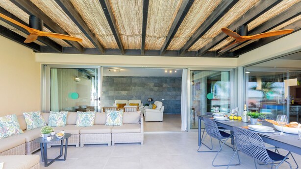 Una sala de estar moderna con un sofá, mesa de comedor y sillas. Se ve un área de cocina a través de puertas de vidrio.