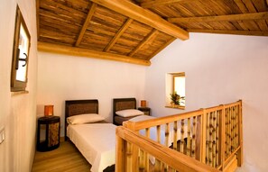 Quarto com duas camas, vigas de madeira no teto, mesa de cabeceira pequena com luminária, espelho e corrimão de madeira.
