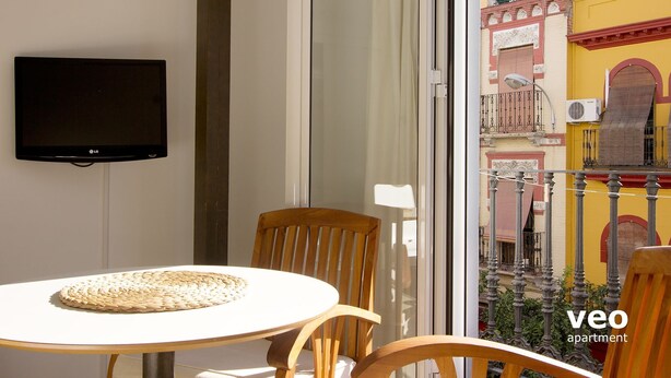 A balcony with a table and chairs, a flat-screen TV mounted on the wall, and a view of a yellow building with balconies. - Studio in Macarena (Sevilla)