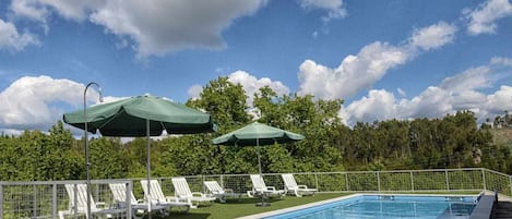 Una piscina con sillas de descanso y sombrillas, rodeada de una valla y árboles.