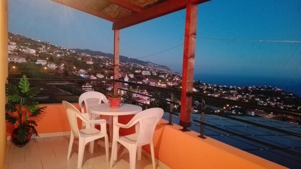 A balcony with a table and chairs, overlooking a hillside town at dusk. - Villa B Apartments (Ponta do Sol)