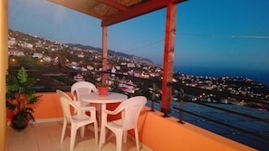 A balcony with a table and chairs, overlooking a hillside town at dusk.