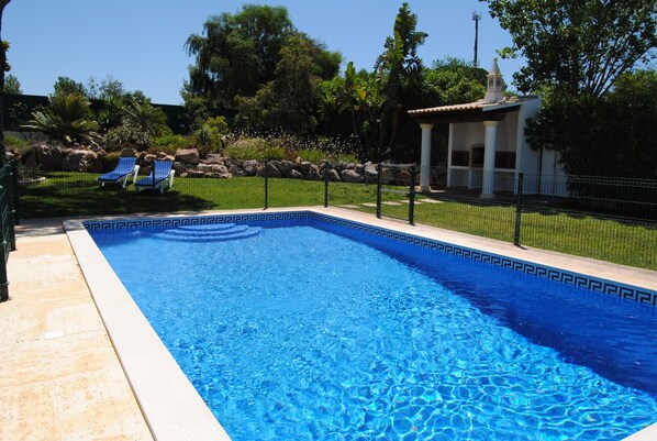 Una piscina con un diseño geométrico de baldosas, rodeada por una valla, con sillas de descanso y un pequeño pabellón al fondo.
