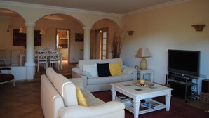 A living room with a white sofa, a coffee table, and a television. There is a dining area in the background with a table and chairs. - Luxury 4 bedroom Villa with private pool and garden (Sta. Eulalia)