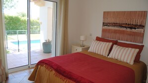A bedroom with a bed, a nightstand, a lamp, a window with a view of a pool, and a painting on the wall. - Luxury 4 bedroom Villa with private pool and garden (Sta. Eulalia)